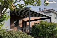 Aluminiowa pergola ogrodowa Square z tkaniną przeciwsłoneczną i punktowym oświetleniem LED na wieczór, montowana przy ścianie domu z cegły, zapewniająca komfortowe miejsce do siedzenia na tarasie z widokiem na ogród