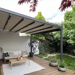 Stylowa pergola ogrodowa Aura z podświetleniem LED i wygodną korbą do sterowania tkaniną z lewej strony, osłaniająca kanapę, fotel i stolik na drewnianym tarasie w otoczeniu zieleni