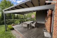 Nowoczesna pergola Square z boczną roletą zewnętrzną ZIP na aluminiowych prowadnicach i w kasecie dopasowanej kolorystycznie do konstrukcji, zamontowana nad tarasem z kwiatami i stołem ogrodowym do ściany domu z cegły z podporą zakotwioną poza tarasem.