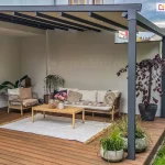 Pergola tarasowa Aura z aluminiową konstrukcją w kolorze antracytowym, ręcznie sterowaną tkaniną i dwoma podporami zamocowanymi do drewnianego tarasu, przytwierdzona do ściany domu nad wnęką tarasową przy ścianie działowej