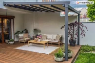 Pergola tarasowa Aura z aluminiową konstrukcją w kolorze antracytowym, ręcznie sterowaną tkaniną i dwoma podporami zamocowanymi do drewnianego tarasu, przytwierdzona do ściany domu nad wnęką tarasową przy ścianie działowej