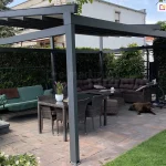 Nowoczesna pergola Square ze skośnym dachem tkaninowym i konstrukcją w kolorze grafitowym w wersji wolnostojącej na tarasie z jacuzzi, huśtawką ogrodową, dużym narożnikiem z poduszkami i stołem jadalnianym, osłaniająca przestrzeń wypoczynkową przed słońcem.