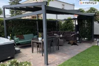 Nowoczesna pergola Square ze skośnym dachem tkaninowym i konstrukcją w kolorze grafitowym w wersji wolnostojącej na tarasie z jacuzzi, huśtawką ogrodową, dużym narożnikiem z poduszkami i stołem jadalnianym, osłaniająca przestrzeń wypoczynkową przed słońcem.