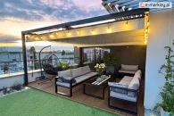 Nowoczesna pergola tarasowa z zamontowanym dachem rzymskim Supro i dekoracyjnym podświetleniem girlandowym – efektowne zadaszenie wypoczynkowej strefy na tarasie z widokiem na miasto, zapewniające ochronę przed słońcem i nadające przytulny klimat.