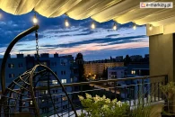 Stylowa roleta rzymska Supro nad nowoczesnym balkonem w zabudowie miejskiej, z efektownym oświetleniem wieczornym i widokiem na osiedle