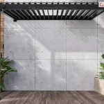 Mała pergola bioklimatyczna Gloria z obracanymi lamelami w kolorze antracytowym, montowana przy ścianie budynku. Konstrukcja wykonana z trwałego aluminium, wyposażona w ręczną korbkę do precyzyjnego sterowania kątem nachylenia lameli dachowych. Otoczenie stanowią donice z roślinami i betonowa ściana, co podkreśla nowoczesny charakter przestrzeni.