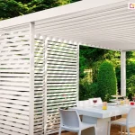 Biała pergola Gloria z aluminiowymi panelami żaluzjowymi zamontowanymi w narożniku. Estetyczne wykończenie konstrukcji tarasowej zapewniające ochronę i elegancję.