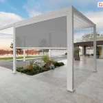 Nowoczesna biała pergola na dużym kamiennym tarasie z opuszczoną szarą roletą screen, widok na basen i jezioro oraz współczesny dom z drewnianą elewacją w tle.