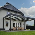 Wypoczynkowa część tarasu ze szklanym zadaszeniem Spekta Glas w kolorze antracytowym postawionym przy białym ceglanym domu z obszernym trawnikiem i kwiatami w drewnianych donicach