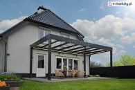 Wypoczynkowa część tarasu ze szklanym zadaszeniem Spekta Glas w kolorze antracytowym postawionym przy białym ceglanym domu z obszernym trawnikiem i kwiatami w drewnianych donicach