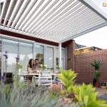 Nowoczesna aluminiowa pergola bioklimatyczna Gloria w kolorze białym zamontowana przy tarasie domu jednorodzinnego. Para siedzi przy stole pod otwartym dachem z obrotowymi lamelami, ciesząc się komfortem cienia i wentylacji. Przestrzeń tarasowa zaaranżowana w stylu ogrodowym z kwiatami i roślinami doniczkowymi. Idealne miejsce do wypoczynku i spożywania posiłków na świeżym powietrzu.