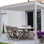 Biała pergola Gloria z zadaszeniem lamelowym nad niewielkim przydomowym tarasem ze stolikiem i kwiatami w doniczkach