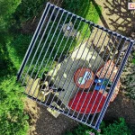 Nowoczesna pergola bioklimatyczna Gloria w wersji wolnostojącej, prezentowana z lotu ptaka, zamontowana w ogrodzie z gęstą roślinnością. Konstrukcja aluminiowa w kolorze antracytowym z dachem z regulowanych lameli zapewnia ochronę przed słońcem i deszczem. Pod pergolą znajduje się elegancka strefa relaksu z dywanem zewnętrznym, ozdobnymi pufami i stolikiem kawowym