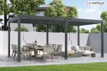 Stylowa pergola aluminiowa Marlo w kolorze grafitowym z lamelowym zadaszeniem. Konstrukcja wolnostojąca, ustawiona na kamiennej posadzce przy białym murze, w otoczeniu zielonego trawnika i drzew. Pod pergolą znajduje się duży, drewniany stół z krzesłami oraz wygodna sofa z poduszkami.