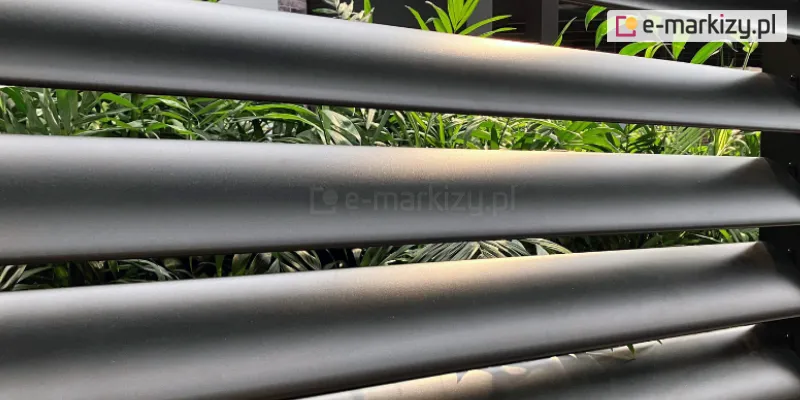 Aluminiowa osłona tarasowa z paneli żaluzjowych Louvre z poziomymi obracanymi lamelami, przez które przebija światło i widoczna jest bujna roślinność ogrodowa w tle. Idealne rozwiązanie przeciwsłoneczne i osłaniające przestrzeń tarasu lub pergoli.