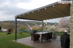 Pergola tarasowa Elegancy Tarasola z aluminiową konstrukcją na dwóch podporach i tkaninowym dachem tworzy komfortową strefę jadalnianą na dużym tarasie. Jasna tkanina dodaje lekkości aranżacji, a w tle rozciąga się widok na zielone pagórki, ogród oraz zabudowania w oddali.