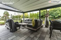 Pergola Elegancy Tarasola z rozciąganym dachem tkaninowym osłania przestrzeń wypoczynkową z wygodnymi fotelami i sofami w nowoczesnym, grafitowym stylu. Zadaszenie idealnie komponuje się z dużym tarasem wyłożonym płytami, otoczonym ogrodem pełnym zieleni.