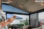 Pergola Elegancy Tarasola z elektrycznie rozsuwanym dachem i zintegrowanym oświetleniem LED tworzy przyjemną, wieczorną atmosferę na tarasie. Na pierwszym planie osoba obsługuje pergolę za pomocą pilota, a w tle widoczny jest ogród oraz nowoczesne meble tarasowe.