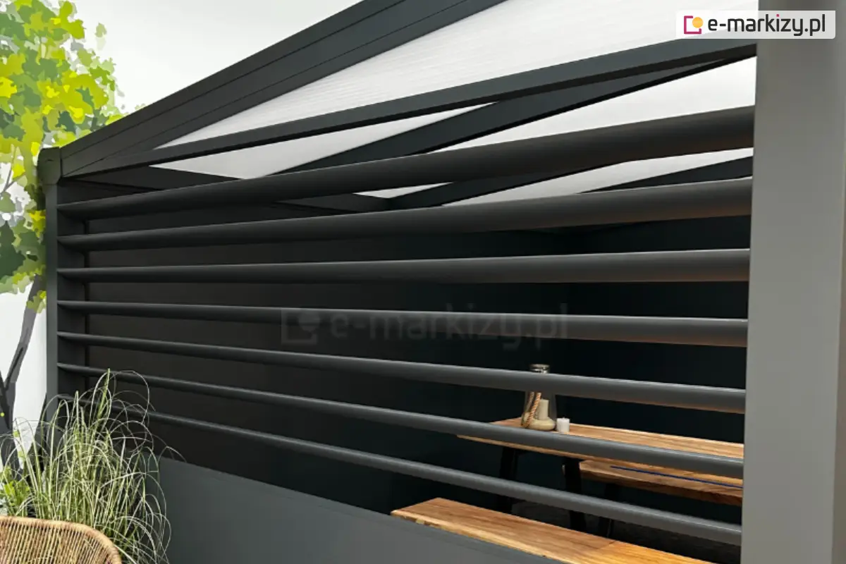 Zewnętrzny, boczny panel żaluzjowy Louvre z poziomymi, ręcznie obracanymi lamelami zamontowany do konstrukcji pergoli ogrodowej; za panelem drewniany stół w przestrzeni jadalnianej, obok zielona roślinność i ściana budynku.