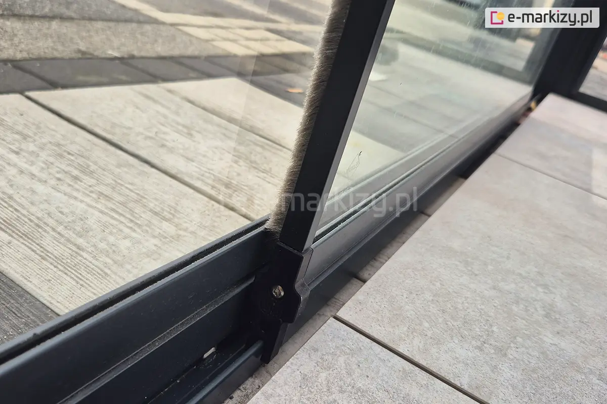 Zbliżenie na system prowadnicy dolnej w zabudowie tarasu Miros z przesuwanymi drzwiami szklanymi. Widoczny profil aluminiowy z osadzonym szkłem oraz szczotką przeciwwietrzną. System zapewnia płynne i łatwe przesuwanie paneli drzwiowych.