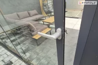 Aluminiowe drzwi szklane systemu Auris z klamką i zamkiem na klucz, zamontowane w całorocznej zabudowie tarasowej. W tle widoczny zestaw wypoczynkowy z sofą narożną i stolikami na kafelkowanym tarasie, otoczonym przesuwnymi panelami szklanymi.