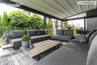 Aluminiowa pergola Elegancy Tarasola z rozciąganym elektrycznie dachem tkaninowym tworzy przestronną, nowoczesną strefę wypoczynkową z kompletem grafitowych sof i stolików. Konstrukcja osłania taras od góry i boków, zapewniając przytulny klimat w otoczeniu zadbanego ogrodu z bujną zielenią.