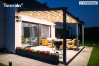 Pergola Elegancy Tarasola prezentuje system LED rozmieszczony w dachu tkaninowym, który podkreśla nowoczesny styl zadaszenia tarasowego. Oświetlona konstrukcja otacza strefę wypoczynku z drewnianymi meblami, a tło stanowi przestronny trawnik.