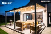 Taras z pergolą Elegancy Tarasola na trzech nogach aluminiowych, wyposażoną w równomierne oświetlenie punktowe LED, tworzy przyjemną, wieczorną strefę do relaksu przy drewnianych meblach ogrodowych. Wokół rozciąga się trawnik oraz spokojna zabudowa jednorodzinna.