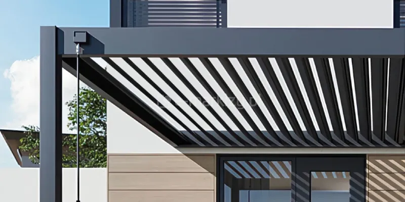 ergola Rubio Soft z ręcznie otwieranym dachem lamelowym pokazuje mechanizm regulacji światła za pomocą ergonomicznej korby. Lamele ustawione pod kątem tworzą dynamiczny rytm cieni, podkreślając minimalistyczną formę pergoli. Konstrukcja przylega do nowoczesnej elewacji, zapewniając skuteczne zadaszenie tarasu i elegancką ochronę ogrodu.