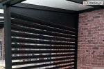 Aluminiowy panel żaluzjowy Louvre w wersji poziomej, zamontowany z boku pergoli tarasowej, skutecznie osłaniający przestrzeń wypoczynkową przed wiatrem i niepożądanymi spojrzeniami, na tle ceglanej elewacji budynku.