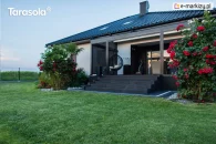 Pergola Elegancy Tarasola zamontowana nad wejściem do domu tworzy nowoczesne, aluminiowe zadaszenie z tkaninowym dachem, osłaniając strefę wypoczynkową z wiszącym fotelem. Konstrukcja harmonizuje z elewacją i otaczającym ogrodem pełnym roślin oraz krzewów róż.