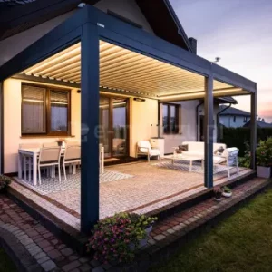 Pergola Essential Tarasola w wersji przyściennej z dachem z obrotowych lameli ustawionych prostopadle do ściany montażowej, sterowanych silnikiem elektrycznym. Konstrukcja w nowoczesnym, głębokim antracycie została zestawiona z jasnymi lamelami, a całość podkreślają nastrojowe punkty LED rozmieszczone pod dachem. Na zdjęciu pergola tworzy zaciszny salon na świeżym powietrzu, otoczony miękkim światłem i wieczorną atmosferą, z kompletem mebli wypoczynkowych ustawionych na kamiennej posadzce tarasu obok domu jednorodzinnego.