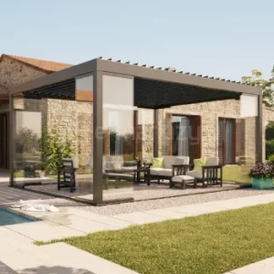 Wolnostojąca pergola Essential Tarasola z lamelowym dachem obracanym elektrycznie tworzy elegancką, nowoczesną strefę wypoczynku na zewnętrznym tarasie. Aluminiowa konstrukcja w ciemnym kolorze subtelnie kontrastuje z jasnymi lamelami, a pełne przeszklenia i rolety zip zapewniają prywatność oraz ochronę przed słońcem. Altanę otacza zadbany ogród oraz kamienna elewacja domu, która nadaje aranżacji śródziemnomorskiego charakteru.