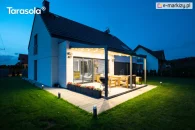 Pergola ogrodowa Elegancy Tarasola z podświetlanym dachem tkaninowym, wsparta na trzech przednich podporach, tworzy stylową przestrzeń wypoczynkową przy domu, z drewnianymi meblami tarasowymi o jasnych poduszkach. Zadaszenie subtelnie rozświetla ogród i sąsiednie budynki w letni wieczór.
