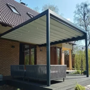 Przyścienna pergola Tarasola Flat o prostokątnej konstrukcji aluminiowej z rozsuwanym dachem tkaninowym i wbudowanym oświetleniem LED osłania elegancką strefę wypoczynkową na tarasie z sofami ogrodowymi. Minimalistyczna forma konstrukcji kontrastuje z ceglaną elewacją domu i zielenią w ogrodzie.