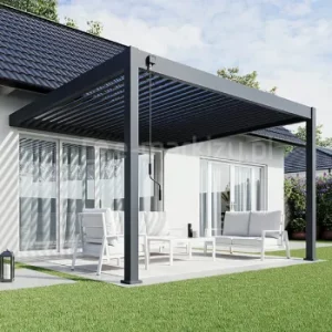 Pergola Rubio Soft z bioklimatycznym dachem lamelowym sterowanym ręczną korbą, z aluminiowymi lamelami ustawionymi w pozycji półotwartej, tworzącymi charakterystyczny cień na tarasie. Konstrukcja w grafitowym kolorze przylega do elewacji budynku, a pod nią znajduje się jasny zestaw wypoczynkowy ustawiony na tarasie otoczonym trawnikiem.