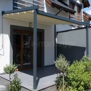 Pergola rzymska Tarasola Simple montowana do ściany, z lekką aluminiową konstrukcją i jasnym dachem tkaninowym składanym w harmonijkę. Osłania niewielki, przydomowy taras z ciemną podłogą oraz roślinnością ogrodową wokół. Smukłe wsporniki i minimalistyczny design podkreślają ekonomiczną formę pergoli.