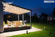 Pergola Elegancy Tarasola z elektrycznie rozsuwanym dachem tkaninowym i intensywnym podświetleniem LED tworzy nastrojowe zadaszenie tarasu z drewnianym kompletem wypoczynkowym. Konstrukcja pięknie rozświetla przestrzeń w otoczeniu zadbanego ogrodu o zmroku.