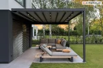 Rozwijana pergola tkaninowa Square Flat firmy Mol z oświetleniem LED w belkach dachowych i rozciągniętą tkaniną w kolorze szarym, zamontowana przy elewacji domu z żaluzjami fasadowymi, osłaniająca narożną sofę oraz leżaki z poduszkami na tarasie ogrodowym wyłożonym płytami gresowymi, otoczonym trawnikiem i zielenią.