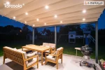 Ścienna pergola rzymska Elegancy Tarasola z podświetlanym dachem tkaninowym tworzy przyjemną, wieczorną strefę wypoczynkową z drewnianymi meblami i grillem. Zadaszenie rozświetla taras, a w tle widoczne są ogród, plac zabaw i zabudowa jednorodzinna.