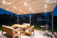 Ścienna pergola rzymska Elegancy Tarasola z podświetlanym dachem tkaninowym tworzy przyjemną, wieczorną strefę wypoczynkową z drewnianymi meblami i grillem. Zadaszenie rozświetla taras, a w tle widoczne są ogród, plac zabaw i zabudowa jednorodzinna.