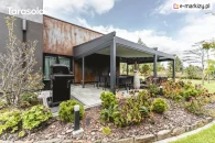 Pergola Elegancy Tarasola z elektrycznie zwijanym dachem i bocznymi roletami tworzy półzamkniętą, elegancką przestrzeń wypoczynkową z technorattanowymi meblami i huśtawką wiszącą. Wnętrze pergoli otwiera się na ogród z widokiem na ogród.