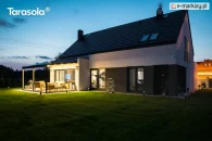 Pergola Elegancy Tarasola z ruchomym zadaszeniem tkaninowym tworzy nowoczesną, oświetloną strefę wypoczynkową na rozległym, zielonym trawniku. Pod pergolą ustawiono drewniany zestaw mebli, a otoczenie budynku podkreśla spokojny, wieczorny klimat.