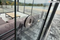 Srebrny uchwyt systemu Auris solidnie zamocowany do szklanego panelu przesuwnego w zabudowie tarasowej z widokiem na jezioro i las. W tle narożna sofa ogrodowa, stolik kawowy i elegancka posadzka z płytek tarasowych.