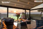 Pergola Elegancy Tarasola z równomiernie rozmieszczonym oświetleniem LED tworzy przytulne wnętrze tarasu wyposażonego w rattanowe sofy, stolik i dekoracje. Przestrzeń otwiera się na ogród z panoramicznym widokiem na pola i altanę ogrodową o zachodzie słońca.
