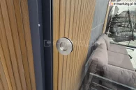 Okrągły uchwyt ze stali nierdzewnej zamontowany na przesuwnym panelu szklanym zabudowy tarasowej Auris. Uchwyt w wersji nielakierowanej, widoczny na tle elewacji z pionowymi deskami oraz narożnika wypoczynkowego w nowoczesnym ogrodzie zimowym.