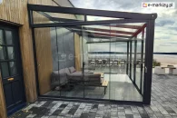 4-metrowa szklana zabudowa tarasowa Auris z panoramicznym widokiem na jezioro. Przesuwne panele szklane chronią strefę relaksu z narożnikiem i stolikiem kawowym przed wiatrem. Konstrukcja aluminiowa przylega do budynku z elewacją drewnianą i kamienną. W tle strefa restauracyjna i plaża.