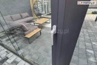 Klamka zamykająca w systemie drzwi przesuwnych Auris zamontowana w aluminiowym profilu, widok na nowoczesne wnętrze zabudowy tarasowej z szarą sofą narożną, stolikiem ogrodowym i fotelem wiszącym, na tle płytek tarasowych i elewacji z drewna.