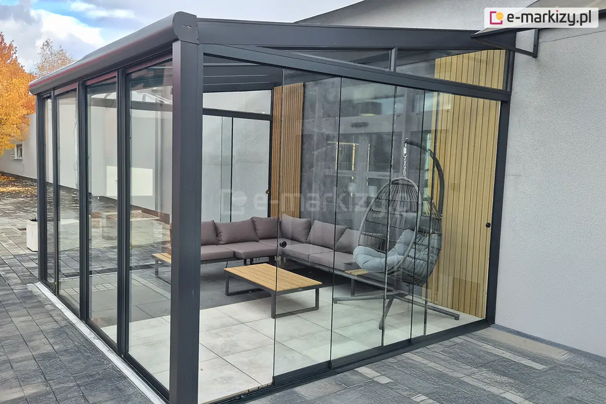 Nowoczesne zsuwane panele szklane Miros zamontowane na tarasie przy elewacji budynku, cztery przesuwne tafle szkła mijające się przy otwieraniu. Zabudowa w aluminiowej ramie w kolorze antracytowym z widocznym narożnym wypoczynkiem i fotelem wiszącym w środku.