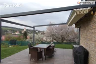 Pergola Elegancy Tarasola z całkowicie zwiniętym dachem rzymskim ułożonym w pakiet, odsłania aluminiową konstrukcję przyścienną nad tarasem z kompletem rattanowych mebli. Pod pergolą znajduje się przestrzeń jadalniana, otoczona ogrodem z kwitnącymi drzewami i widokiem na pobliską zabudowę.